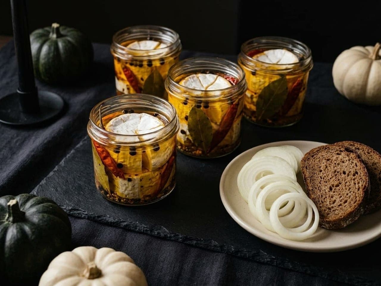 Small clear jars of marinated soft white-rind cheese in golden oil with whole dried red chillies and bay leaves against the glass, fresh onion rings on a pale plate beside them