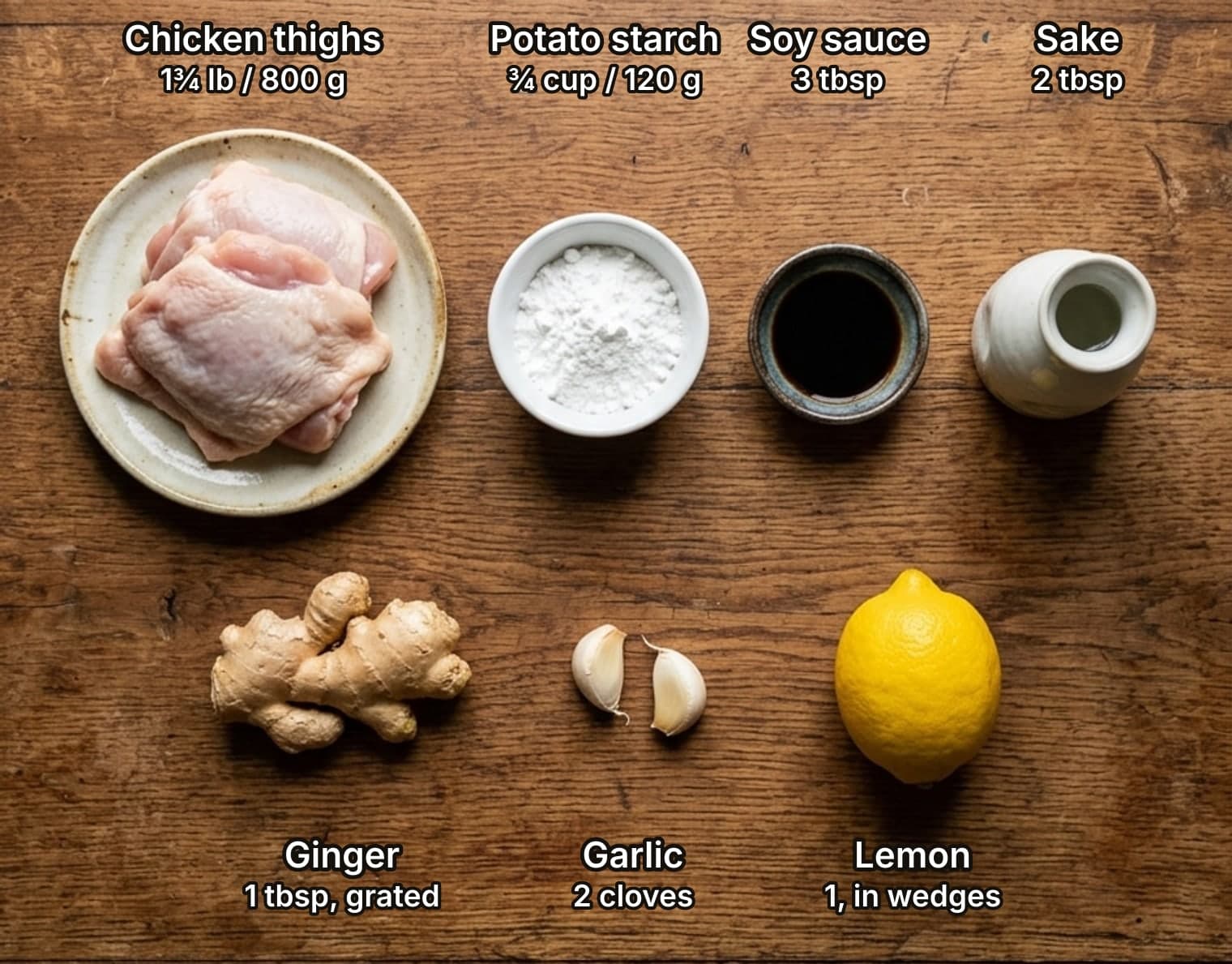 Ingredients for karaage laid out overhead on a wooden kitchen counter and labelled with quantities: chicken thighs on a plate, a bowl of potato starch, a dish of soy sauce and a small flask of sake, with a hand of ginger, two garlic cloves and a lemon below.