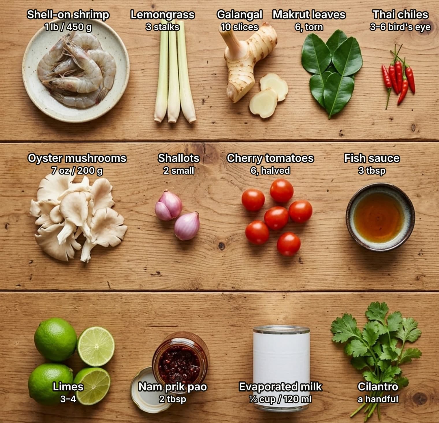 Thirteen tom yum ingredients laid out overhead on a wooden kitchen counter and labelled with quantities: shell-on shrimp, lemongrass, galangal, makrut lime leaves and bird's eye chiles on top; oyster mushrooms, shallots, cherry tomatoes and fish sauce in the middle; limes, nam prik pao chili jam, evaporated milk and cilantro below.