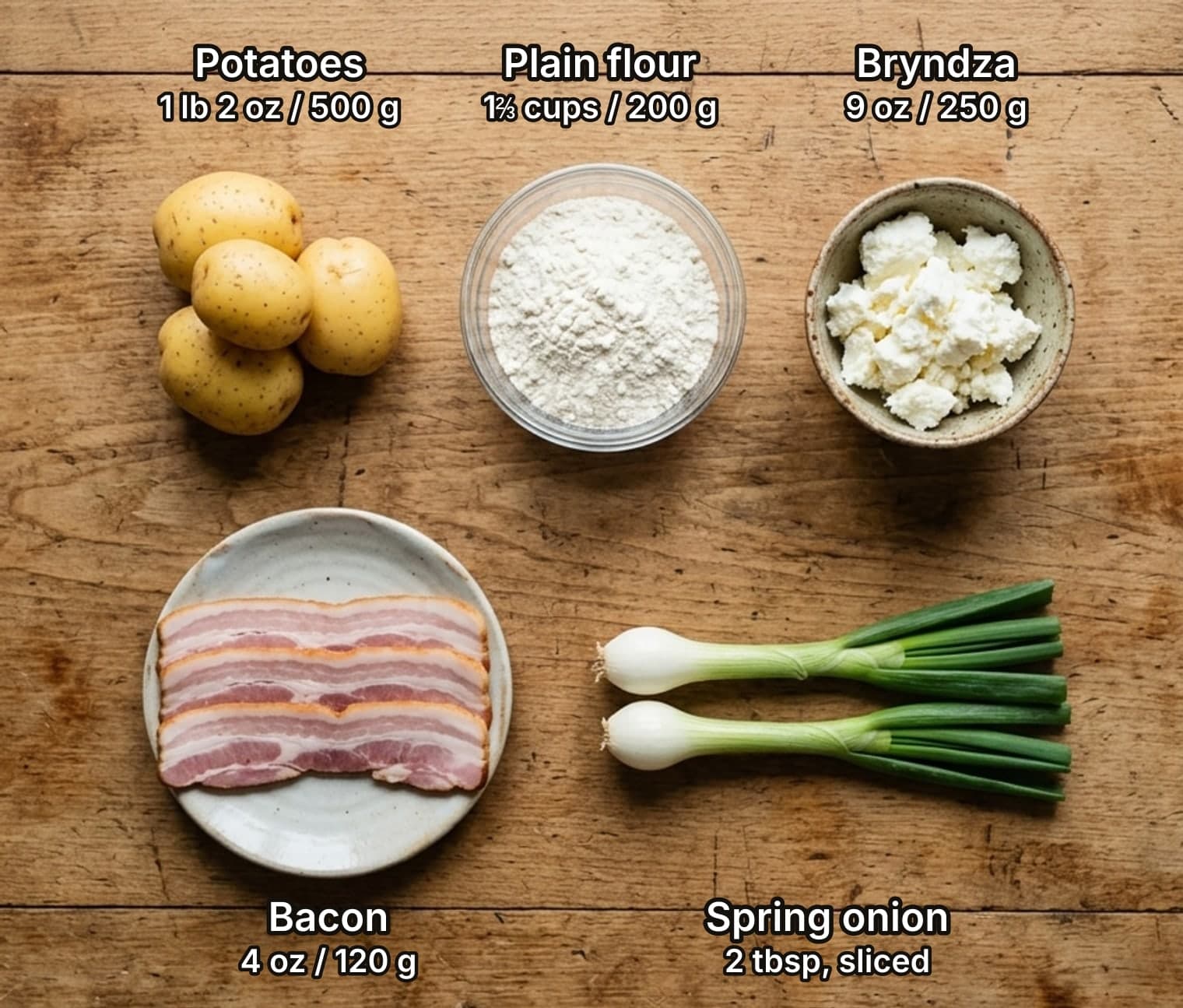 Ingredients for halušky with bryndza laid out overhead on a wooden kitchen counter and labelled with quantities: potatoes, a bowl of plain flour and a bowl of crumbly bryndza sheep cheese for the dumplings, with a plate of bacon strips and two spring onions to finish.