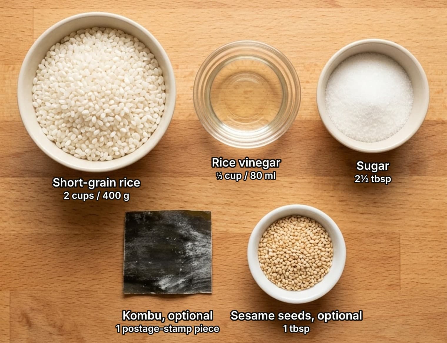 Sushi rice ingredients laid out overhead on a wooden counter and labelled with quantities: a bowl of raw Japanese short-grain rice, a glass dish of rice vinegar and a bowl of sugar across the top; a square of dried kombu and a small bowl of toasted sesame seeds below.