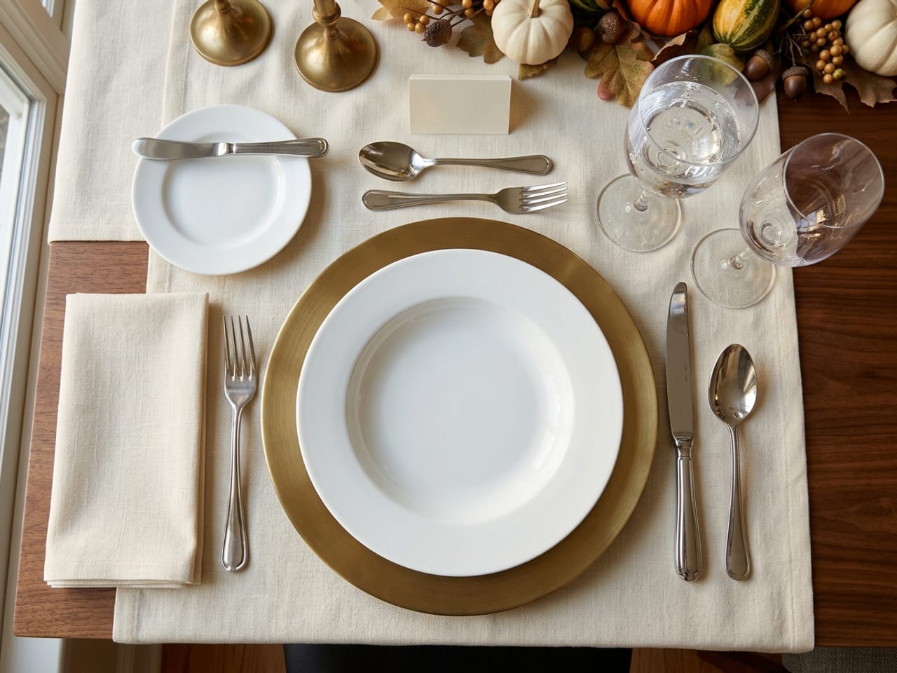 Overhead formal Thanksgiving place setting: a brass charger with a white soup plate, napkin and dinner fork to the left, knife and soup spoon to the right, bread plate with butter knife upper left, dessert spoon and fork above, water and wine glasses upper right, and a blank place card