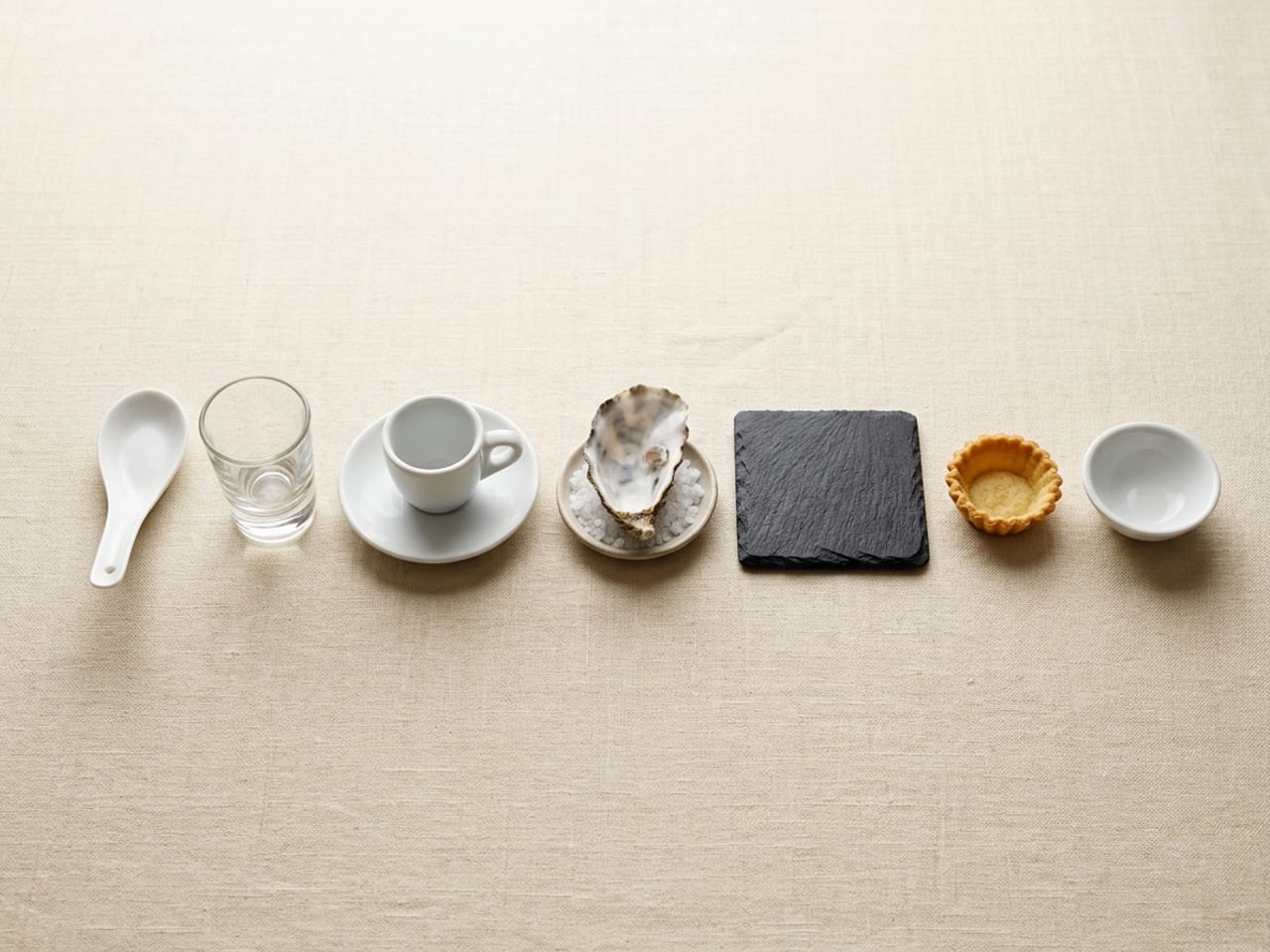 Seven empty amuse-bouche vessels in a row on cream linen: a white Chinese soup spoon, a shot glass, an espresso cup on its saucer, an oyster shell on a dish of coarse salt, a square of dark slate, a small fluted pastry case and a small white bowl