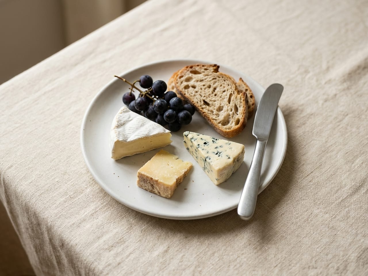 A white dessert plate with small wedges of different cheeses, a few thin slices of bread, a small cluster of grapes and a cheese knife, on cream linen