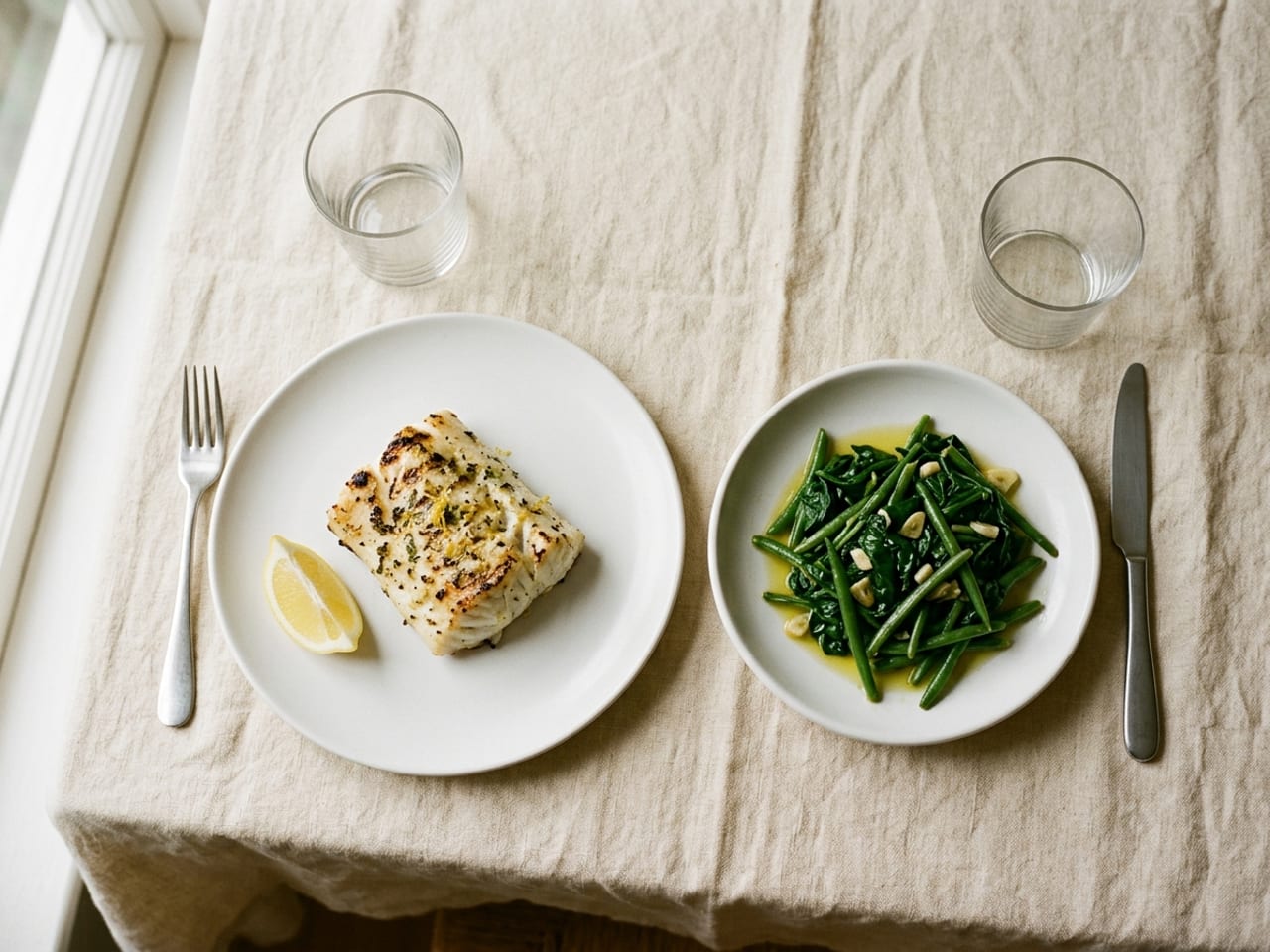 A white dinner plate with a piece of grilled fish and a lemon wedge, and beside it a separate small white dish of sautéed green vegetables, on cream linen
