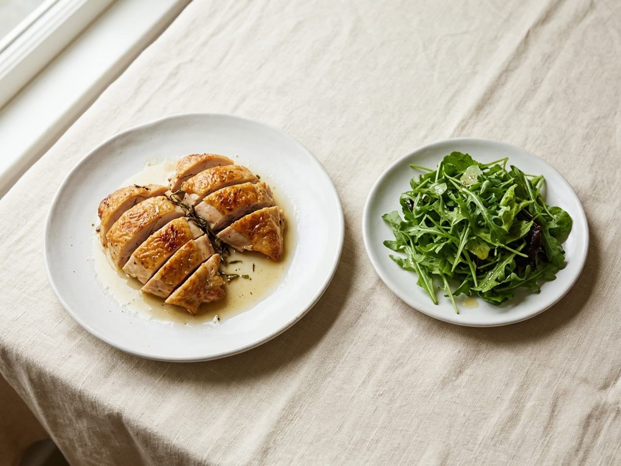 A white dinner plate with slices of roast chicken and a little clear juice, and beside it on the same cream linen a small white plate of plain dressed green salad leaves