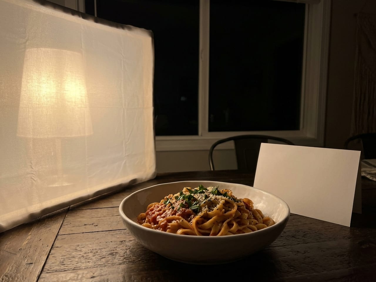 AI illustration of one lamp at night: a bowl of pasta on a dark table in a dim room, lit from the left by a single lamp shining through a white diffusing sheet, a white card standing on the right, a dark window behind