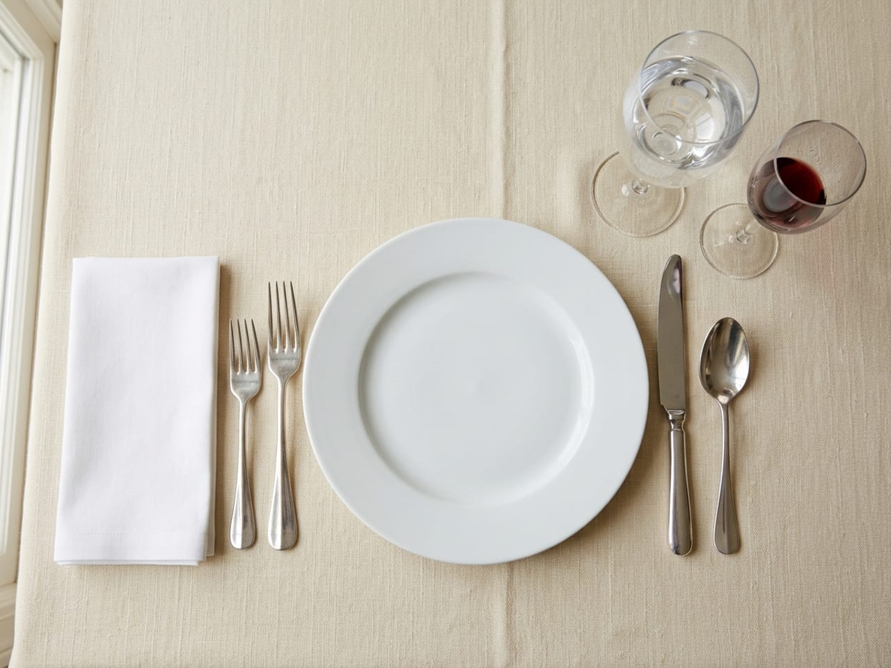 A place setting from above on cream linen: a white dinner plate, two forks on the left with a white napkin folded into a flat oblong beside them, a knife and spoon on the right and a water glass above the knife