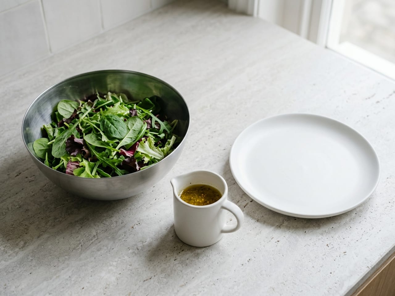 Washed, dry mixed salad leaves in a large stainless mixing bowl on a pale counter, a small white jug of vinaigrette beside the bowl and an empty white plate next to it