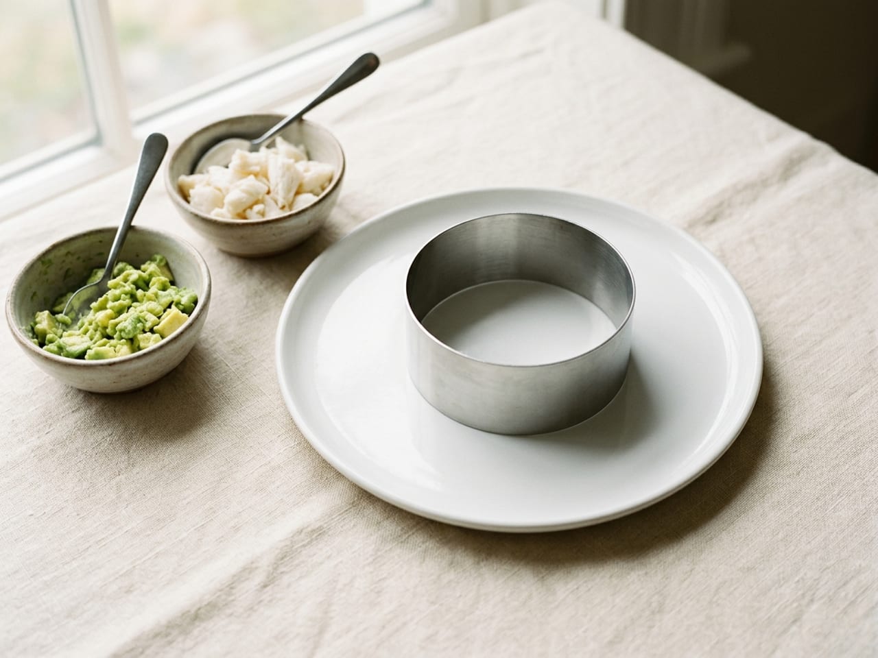 A plain round stainless steel ring mold standing empty on a plain white plate, and beside the plate on the linen a small bowl of diced, lightly mashed avocado and a small bowl of white crab meat, a teaspoon in each