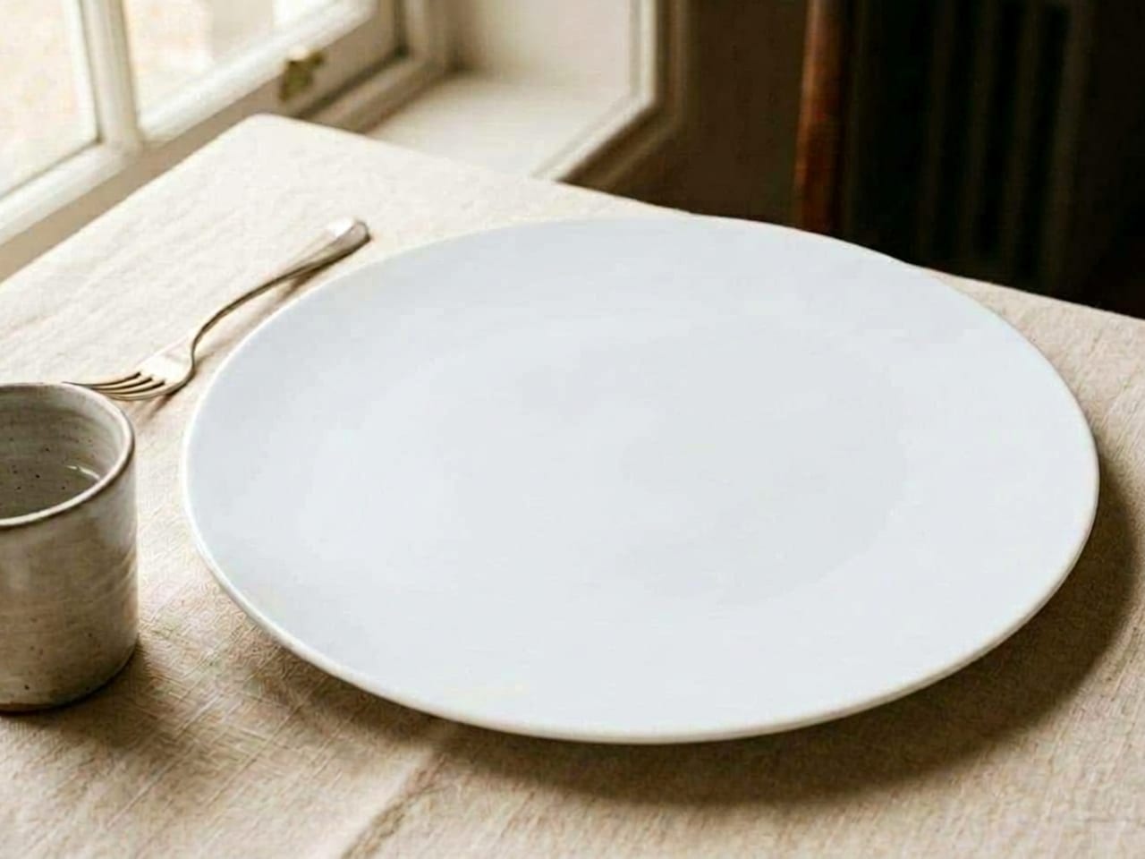 An empty oversized matte white plate on cream linen, a fork and a stoneware cup beside it