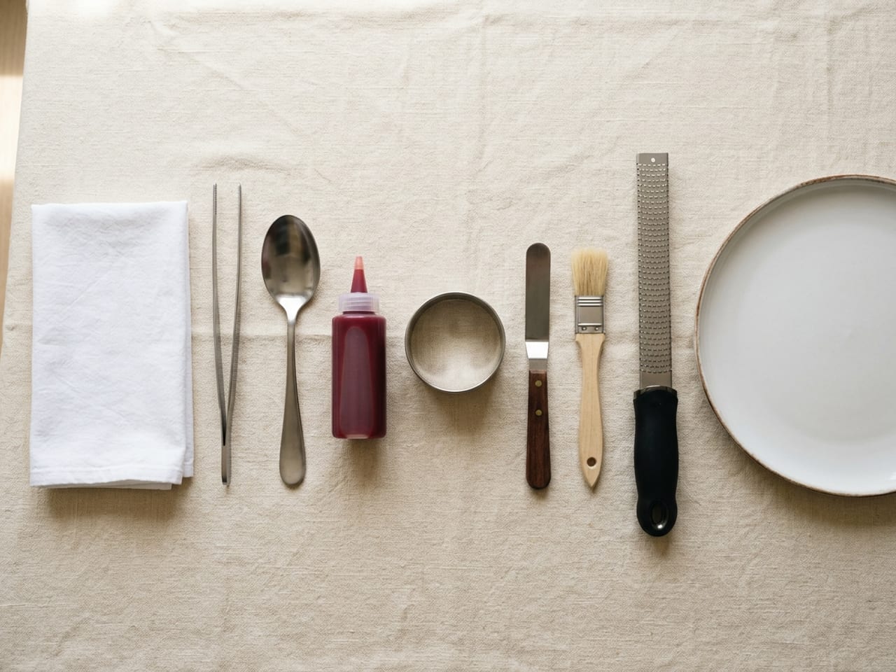 A plating kit laid out in a neat row on cream linen beside an empty matte white plate: a folded white cloth, a pair of steel tweezers, a large steel spoon, a clear plastic squeeze bottle of red sauce, a steel ring, a small offset spatula, a pastry brush and a long narrow rasp grater
