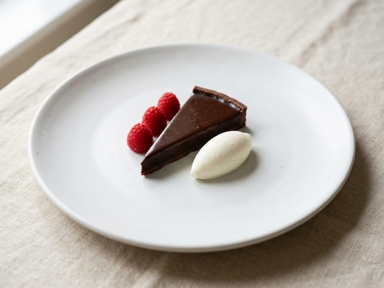 A thin slice of glossy dark chocolate tart on a matte white coupe plate with a small quenelle of crème fraîche and three fresh raspberries