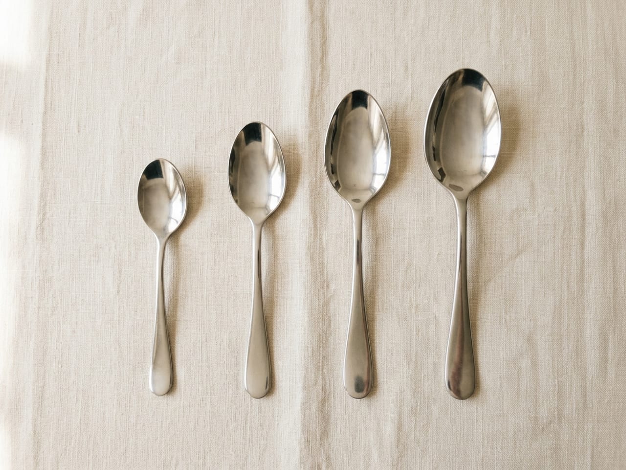 Plain silver spoons of increasing size laid side by side on cream linen, bowls up, from a small coffee spoon on the left to a large serving spoon on the right, an oval dessert spoon with a deep bowl among them