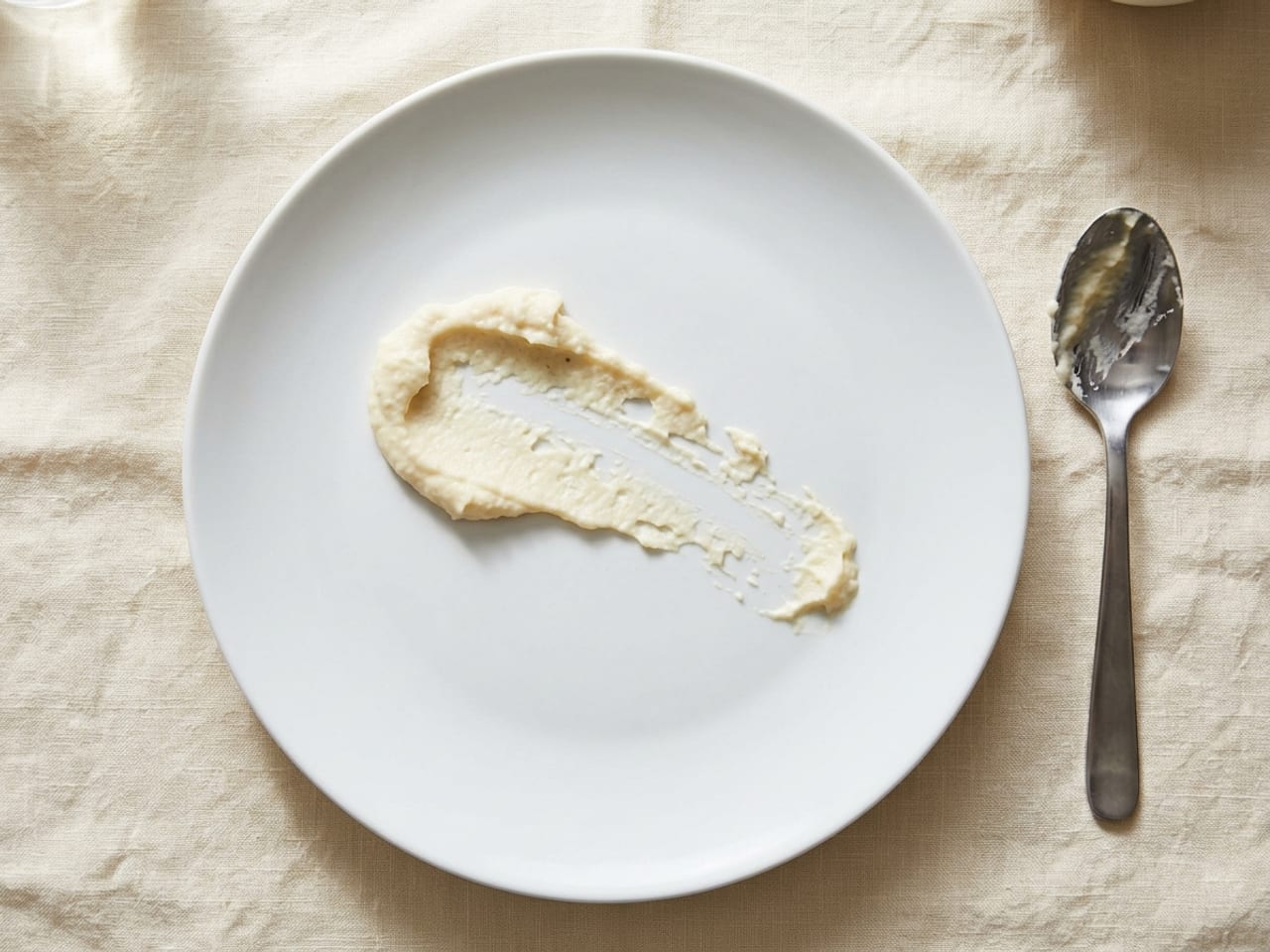 Overhead view of a matte white plate with a failed swoosh of thick ivory purée: the mark is patchy and broken along its length, with ragged edges and gaps of bare plate, a spoon lying beside it