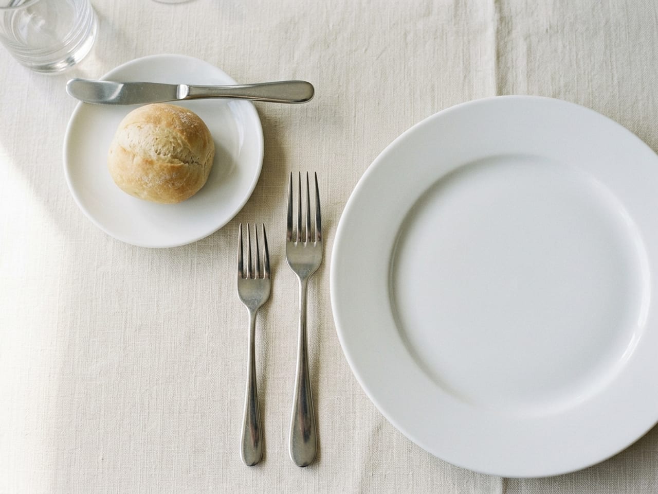 The left half of a place setting from above: two forks beside a white dinner plate, and above the forks a small bread plate holding a roll, with a butter spreader across its top edge, handle to the right