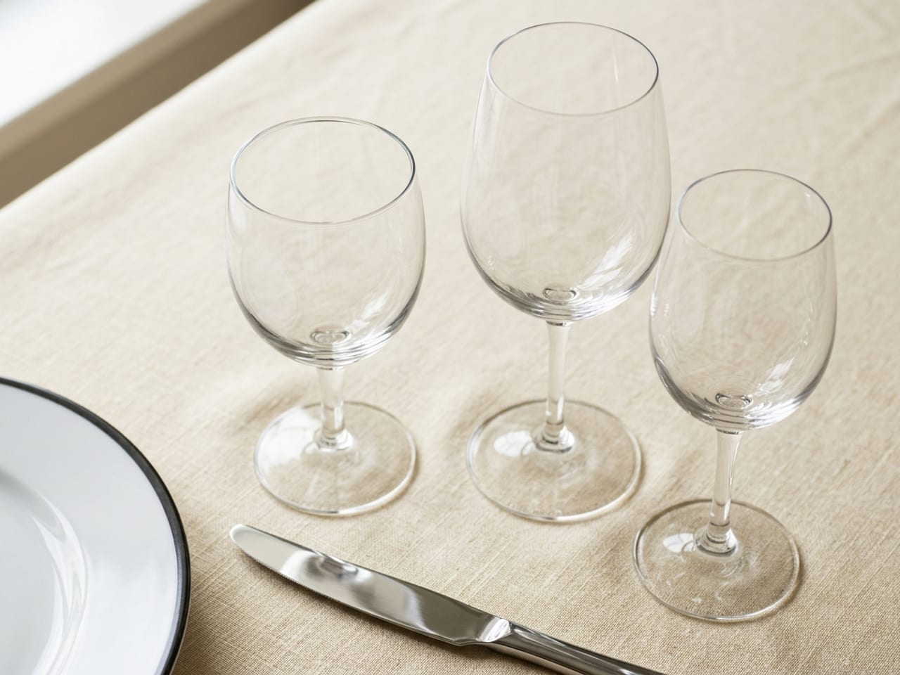 Close on the upper right of a formal place setting: the tip of a silver dinner knife at the bottom, a water goblet just above it, and a red wine glass and a smaller white wine glass stepping down to the right in a gentle diagonal