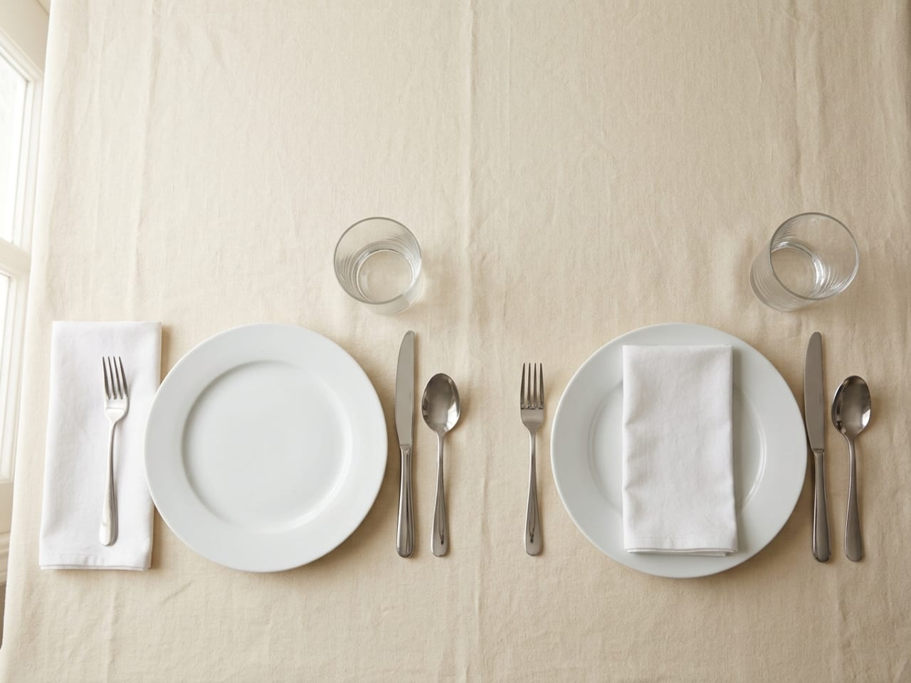 Two simple place settings side by side from above: on the left a white napkin folded into an oblong lies left of the plate with the fork resting on it; on the right the same napkin lies folded flat on the centre of the plate, the fork beside it on the cloth