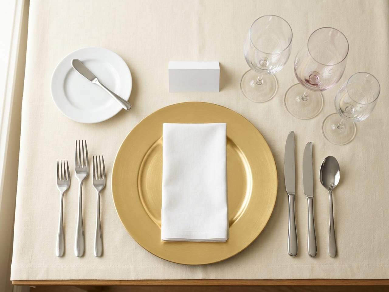 A formal place setting from directly above: a gold charger with a white napkin folded on it, three forks on the left (fish, dinner, salad), a dinner knife, fish knife and soup spoon on the right, a bread plate with a butter spreader at the upper left, a blank place card above the charger, and a water goblet, red wine glass and white wine glass in a short diagonal at the upper right