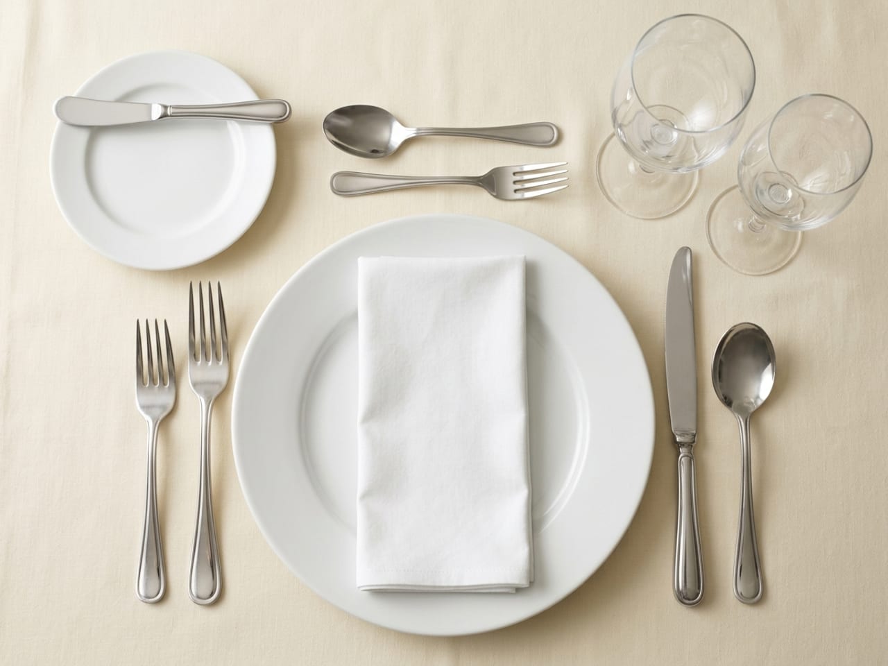 An informal dinner-party place setting from directly above: a white dinner plate with a folded napkin on it, a salad fork and a dinner fork on the left, a knife and a soup spoon on the right, a small bread plate with a butter spreader at the upper left, a dessert spoon and fork lying across the top of the plate, and a water goblet and a wine glass at the upper right