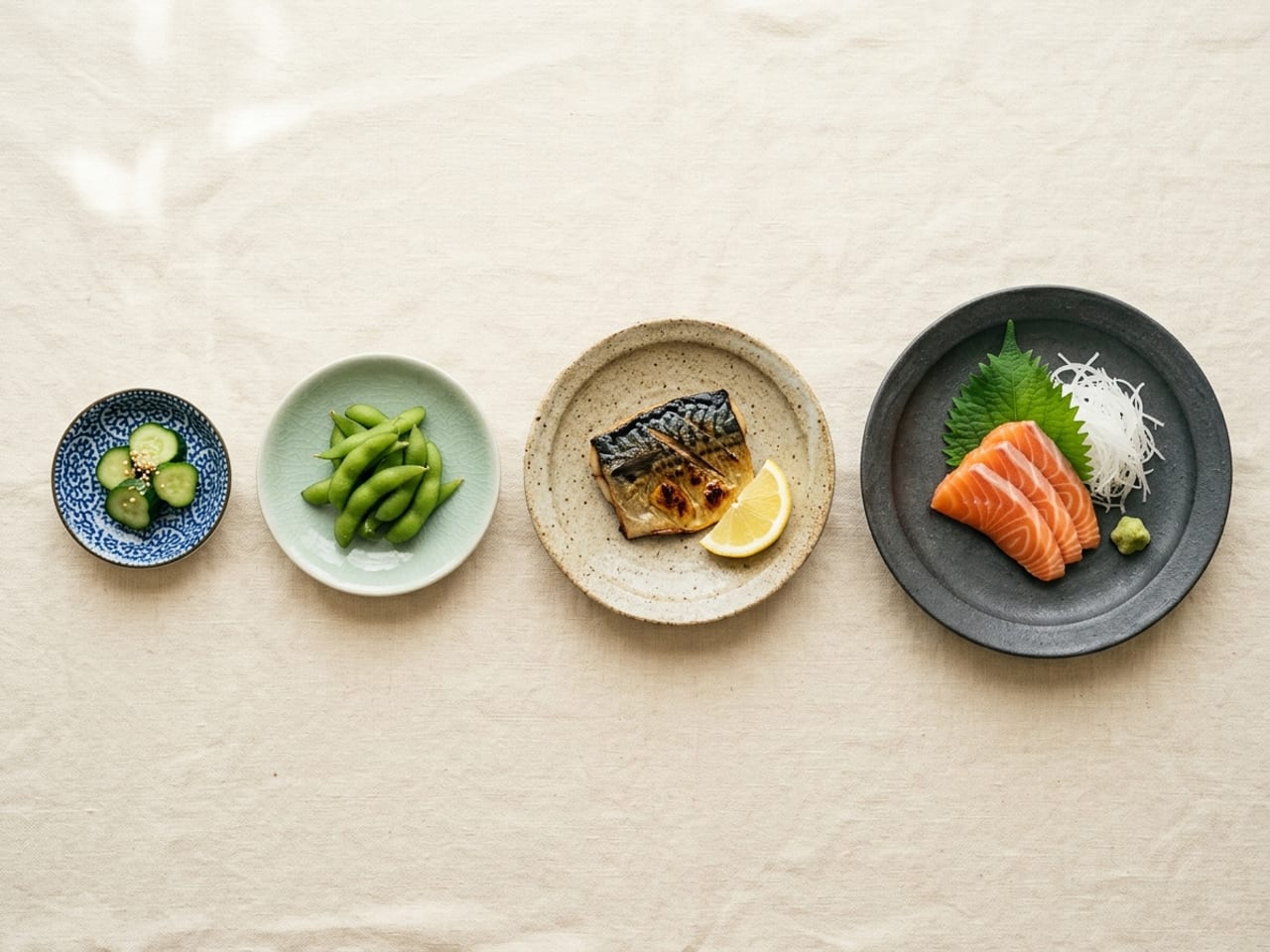 Four Japanese ceramic plates in a row from small to large on cream linen: a tiny indigo-patterned mamezara with pickled cucumber slices, a pale celadon kozara with edamame, a rustic oatmeal chūzara with grilled mackerel and lemon, and a large matte charcoal ōzara with salmon sashimi, daikon and a shiso leaf