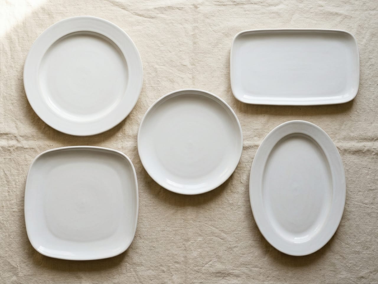 Five empty white porcelain plates on cream linen in a loose grid, each a different shape: a round plate with a wide flat rim, a round rimless coupe with gently upturned edges, a square plate with softened corners, a long rectangular plate and an oval plate