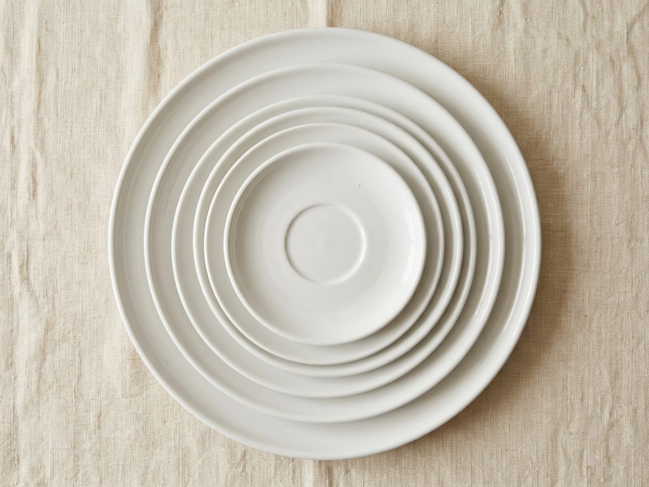Plain white porcelain plates nested concentrically on cream linen, each centred on the next larger one so every rim shows as a ring, from a large charger at the bottom up to a saucer on top