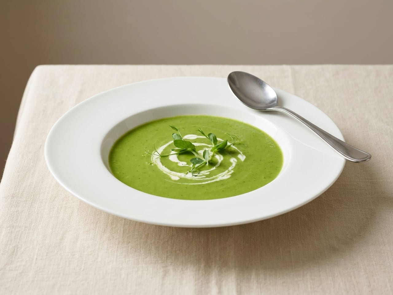 Green pea soup with a swirl of cream and pea shoots in the deep well of a white soup plate with a broad flat rim, a spoon resting on the clean rim