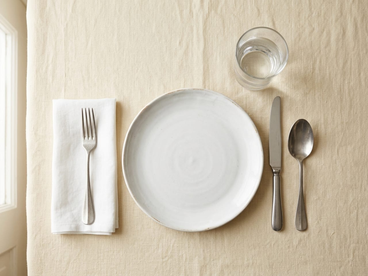 A basic place setting seen from above: a white dinner plate, a fork on a folded napkin to the left, a knife and soup spoon to the right and a water glass above the knife